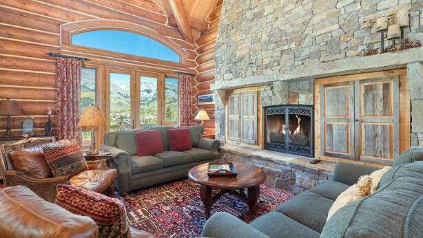 A log cabin interior with a stone fireplace, leather furniture, and a large window overlooking a mountainous landscape. - Townhomes On The Creek 119 (Mountain Village)