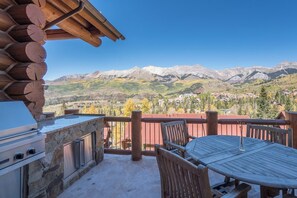 A wooden deck with a stone barbecue, a table, and chairs, overlooking a mountainous landscape. - Townhomes On The Creek 119 (Mountain Village)