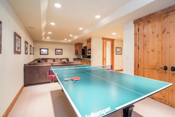 A ping pong table in a room with a sofa, wooden door, and framed pictures on the wall.