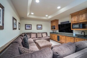 A living room with a sectional sofa, a flat-screen TV, wooden cabinets, and a kitchen area with a microwave.
