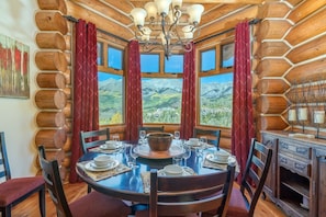 L’intérieur d’une cabane en rondins, avec une table à manger dressée pour quatre personnes, entourée de chaises, et de grandes fenêtres offrant une vue sur les montagnes.
