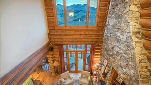A log cabin interior with a stone wall, wooden ceiling, and a view of mountains through large windows. - Villas at Tristant 205 (Mountain Village)