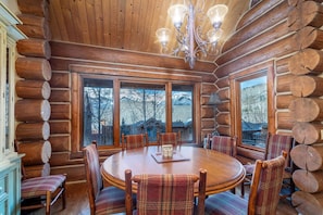 Ein Innenraum einer Blockhütte mit einem runden Holztisch, der von karierten Stühlen umgeben ist, großen Fenstern mit Blick auf eine verschneite Landschaft und einer Kristallleuchtre.