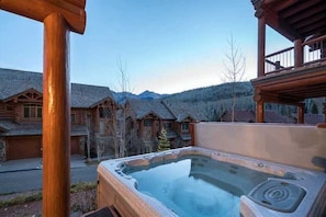 Une cabane en bois avec un jacuzzi à l’avant, entourée d’autres cabanes et avec des montagnes en arrière-plan.