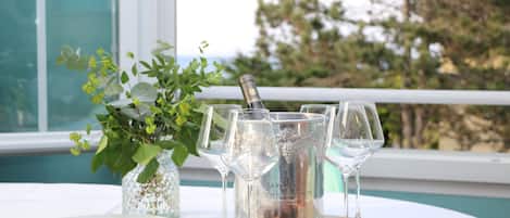 Mesa servida con una botella de champán, copas, una tabla de madera con embutidos y pequeños recipientes de salsas, con vista a árboles y un edificio al fondo.