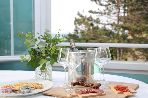 A table set with a bottle of champagne, glasses, a wooden board with sliced meats, and small bowls of dips, with a view of trees and a building in the background.