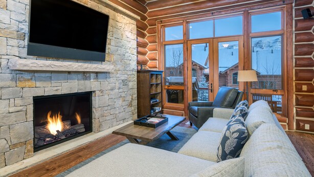 A log cabin interior with a stone fireplace, a large flat-screen TV, a sofa, and a view of a snowy landscape through large windows. - Spacious 4 Bedroom Villa with Ski-in Ski-out Access (Mountain Village)