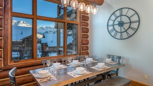 A dining area with a wooden table set for four, a large wall clock, and a window with a view of mountains. - Spacious 4 Bedroom Villa with Ski-in Ski-out Access (Mountain Village)