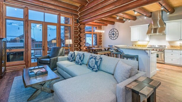 A cozy living room with a sofa, coffee table, and a fireplace. There is a kitchen area with cabinets and a stove. Large windows offer a view of the outside. - Villas At Tristant 201 (Mountain Village)