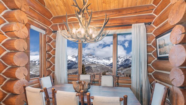 Ein Blockhaus-Interieur mit Esstisch, Stühlen, einer Chandelierleuchtre und Blick auf schneebedeckte Berge.