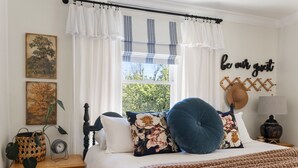 Ein Schlafzimmer mit Bett, Nachttisch, einem Fenster mit Vorhängen, einer Wanduhr, einer Lampe und einem motivierenden Zitat.