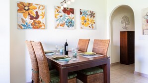 Uma sala de jantar com uma mesa de madeira posta para quatro pessoas, rodeada por cadeiras de vime com almofadas verdes. Acima da mesa, há três quadros florais na parede.