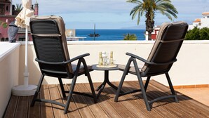 Duas cadeiras com uma mesa no meio, em um deck de madeira, com vista para a água e palmeiras.