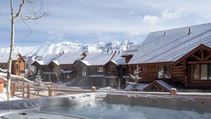 Una cabaña de madera con un jacuzzi en la parte delantera, rodeada de montañas nevadas.
