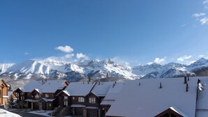 Una hilera de cabañas de madera con techos cubiertos de nieve, frente a un paisaje de montañas nevadas bajo un cielo azul despejado.