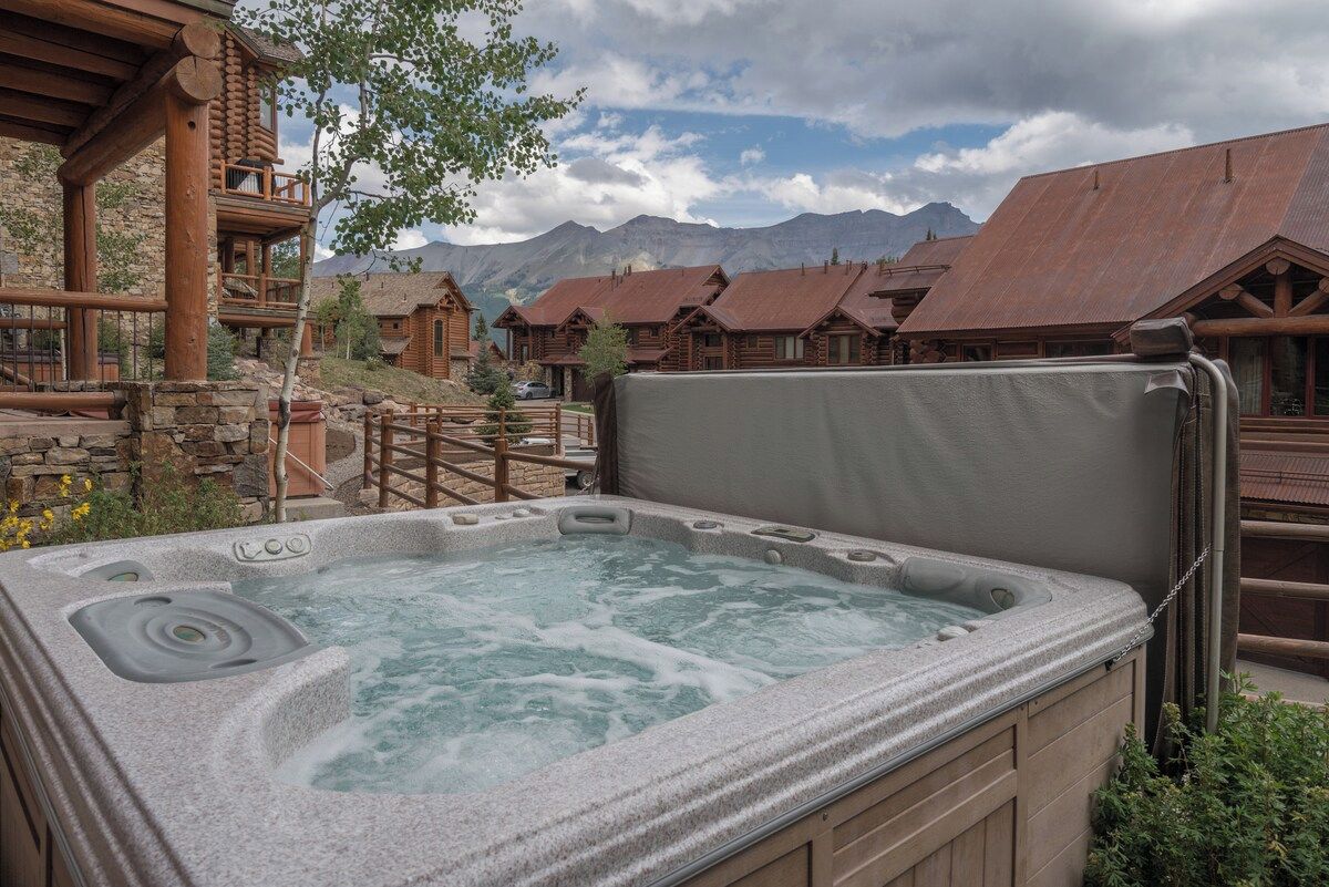 A hot tub with a stone exterior, surrounded by wooden cabins with metal roofs, set against a mountainous backdrop.