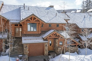 A log cabin with a stone foundation, a wooden garage door, and a snow-covered roof. - Villas At Tristant 221 (Mountain Village)