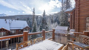 Una terraza de madera con una parrilla y asientos, con vista a un bosque nevado y montañas.