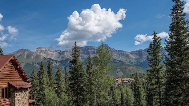 A log cabin with a stone foundation, surrounded by dense evergreen trees and a backdrop of rugged mountains under a clear blue sky. - Villas At Tristant 127 (Mountain Village)