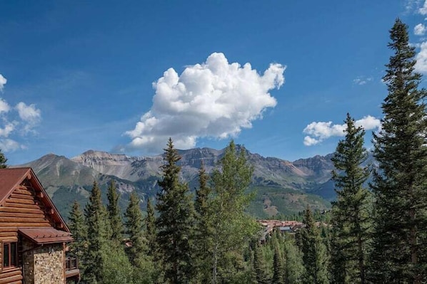 Une cabane en rondins, posée sur des fondations en pierre, entourée d’une forêt dense de conifères et dominée par des montagnes escarpées sous un ciel bleu dégagé.