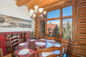 A dining area with a wooden table set for six, surrounded by chairs, with a mountain view through large windows.