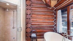 Ein Badezimmer in einer Blockhütte mit einer Duschkabine aus Glas, einer Wand aus Holzstämmen, einer Badewanne und einem Fenster mit Vorhängen.