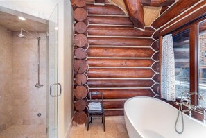 Une salle de bain rustique, aménagée dans une cabane en rondins, avec une douche à parois de verre, un mur en rondins de bois, une baignoire et une fenêtre avec des rideaux.