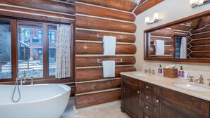 A log cabin bathroom with a large bathtub, wooden walls, a window, and a vanity with a mirror.