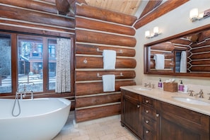 A log cabin bathroom with a large bathtub, wooden walls, a window, and a vanity with a mirror.