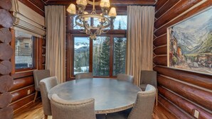 A log cabin interior with a round table and chairs, a chandelier, large windows with curtains, and a painting of a mountain landscape.