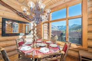 A log cabin interior with a dining table set for four, a chandelier, and a large window offering a mountain view.