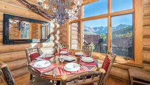 Comedor de cabaña de troncos con un ventanal grande que ofrece vistas a la montaña, una araña y muebles de madera rústica.