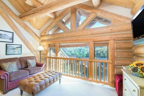 A log cabin interior with a wooden ceiling, a sofa, an ottoman, a TV, and a balcony with a view.