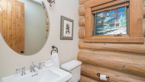 A bathroom with a wooden log wall, a round mirror, a sink with faucets, a toilet, and a window with a view of trees. - Villas At Tristant 215 (Mountain Village)