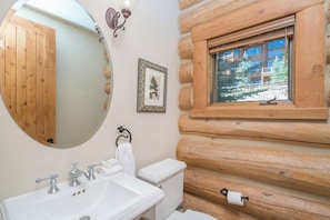 Une salle de bain avec un mur en rondins de bois, un miroir rond, un lavabo avec des robinets, des toilettes et une fenêtre donnant sur les arbres.