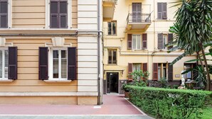 Un edificio de varios pisos con balcones, entrada empedrada en rojo y una estatua en el jardín.