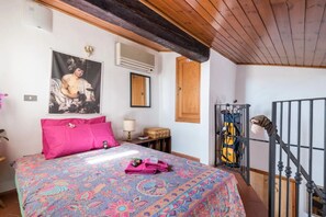 Um quarto com cama, teto de madeira, um pôster na parede, uma mala e uma bicicleta.