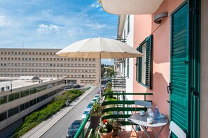 Un balcone con un ombrellone bianco, un tavolo con tazze e una brocca, e vista su una strada con auto e palazzi.