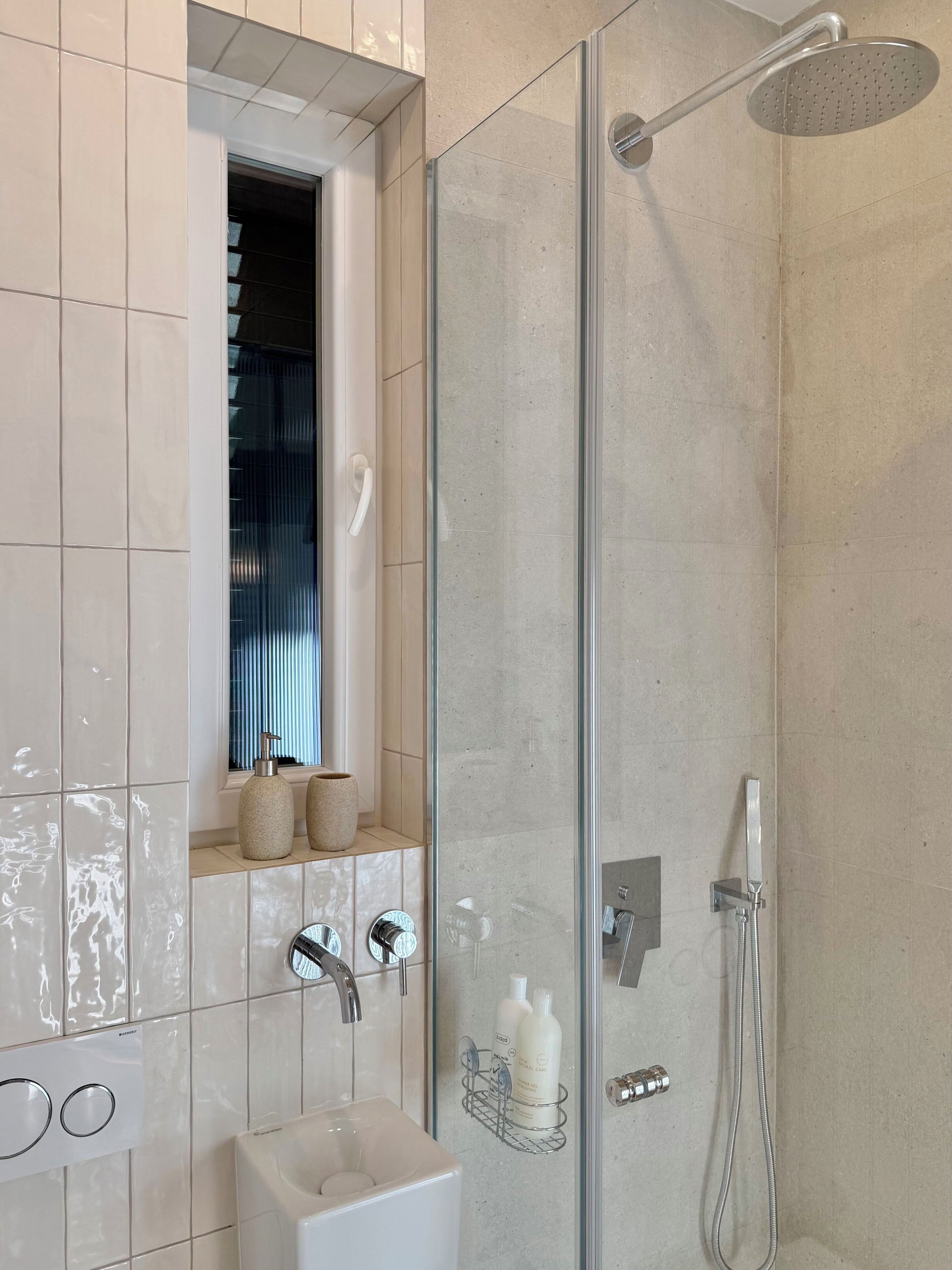 A modern bathroom with a glass-enclosed shower, a sink with a soap dispenser, and a window above the sink.