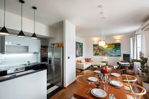 A modern kitchen with stainless steel appliances, a dining area with a wooden table set for four, and a living room with a sofa and artwork.