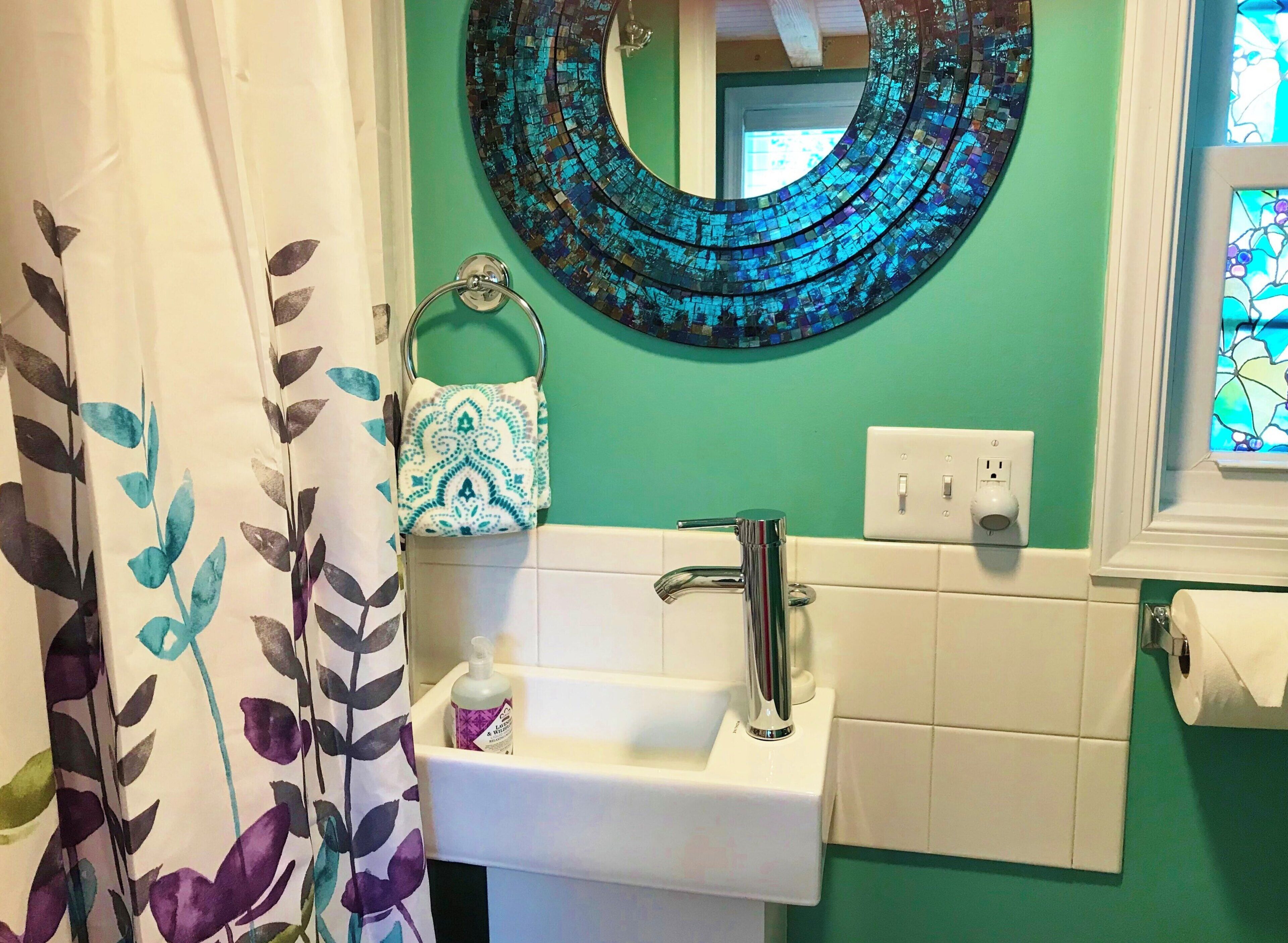Une salle de bain avec un mur vert, un lavabo blanc, un miroir et un rideau de douche orné d’un motif de feuilles.