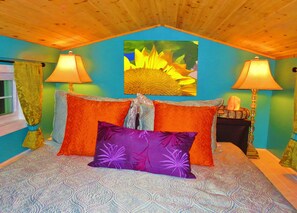 A bedroom with a patterned bedspread, two lamps, a painting of a sunflower, and a window with curtains.