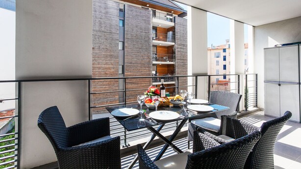 A balcony with wicker furniture, a table set for dining, and a view of a building with balconies. - VICTOR HUGO AP4159 by RIVIERA HOLIDAY HOMES (Niza)