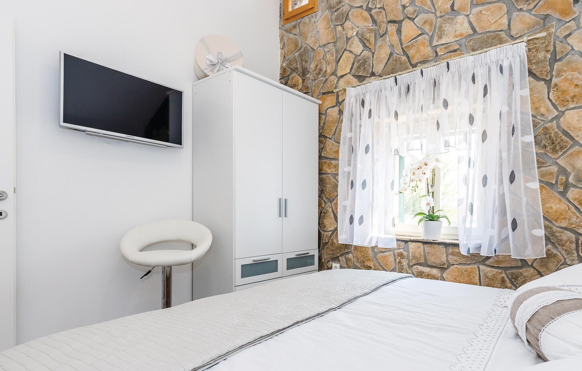 Ein Schlafzimmer mit Steinwand, einem Bett mit weißer Bettwäsche, einem weißen Kleiderschrank, einem kleinen weißen Hocker und einem Fernseher an der Wand.