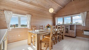 A wooden cabin interior with a dining table, chairs, a kitchen area, and large windows offering a view of the outdoors.