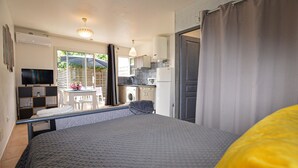 Een slaapkamer met een bed, een televisie, een tafeltje met bloemen, een wasmachine en een keuken met een koelkast en magnetron.