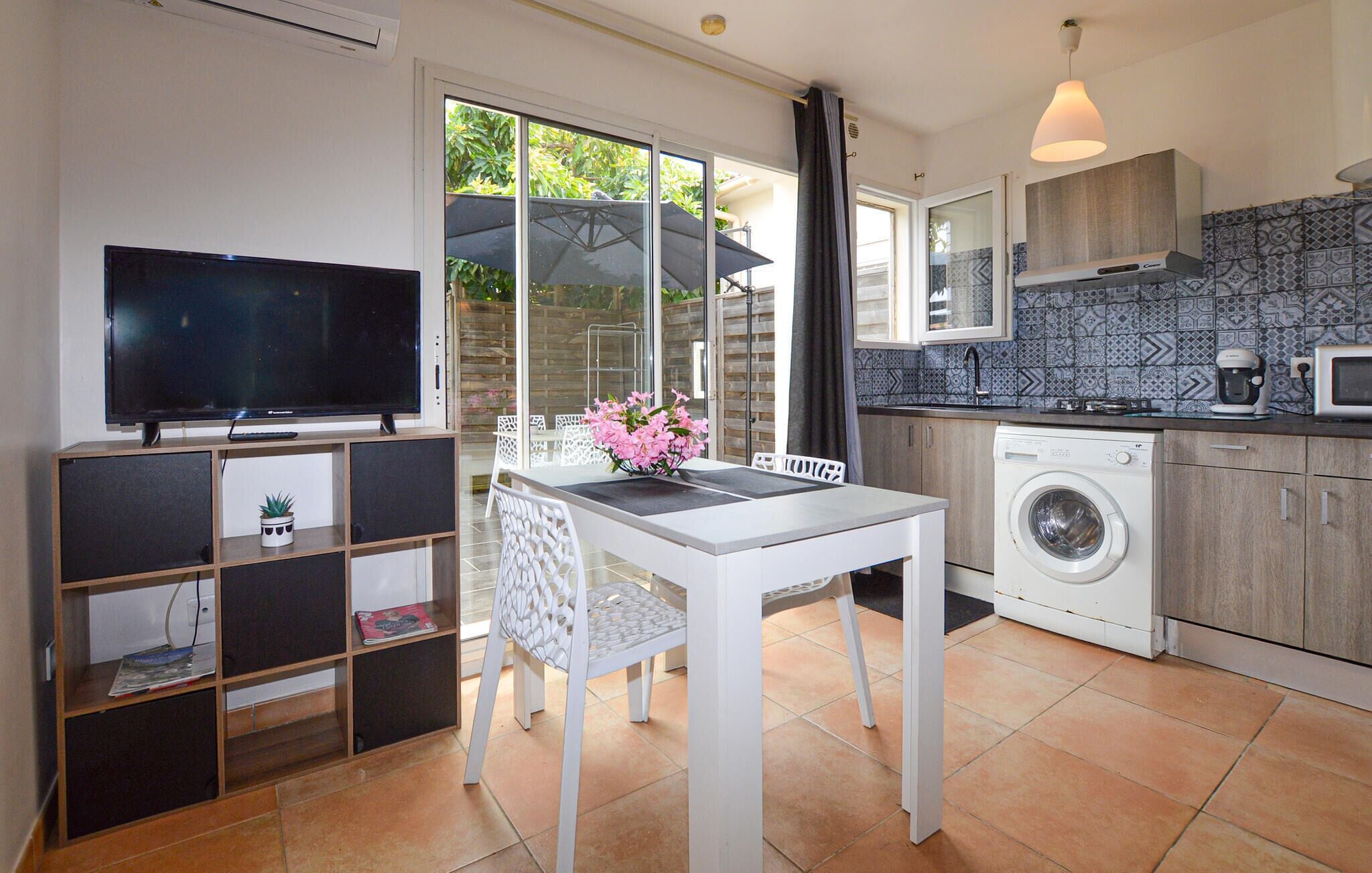 Een kleine keuken met een wasmachine, een eettafel met stoelen, een tv op een dressoir en uitzicht op buiten.