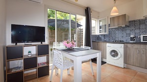 Een kleine keuken met een wasmachine, een eettafel met stoelen, een tv op een dressoir en uitzicht op buiten.