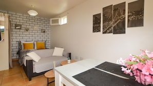 Een slaapkamer met een metselwerk muur, een bed met gele en witte kussens, een klein tafeltje met een plant, en drie ingelijste foto’s aan de muur.