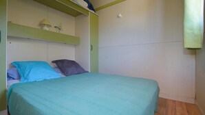 A single bed with a teal bedspread and two pillows, a small shelf with a lamp and a plant, and a white wall with a smoke detector. - 2 bedroom awesome (sta)caravan in Casalabriva (Casalabriva)